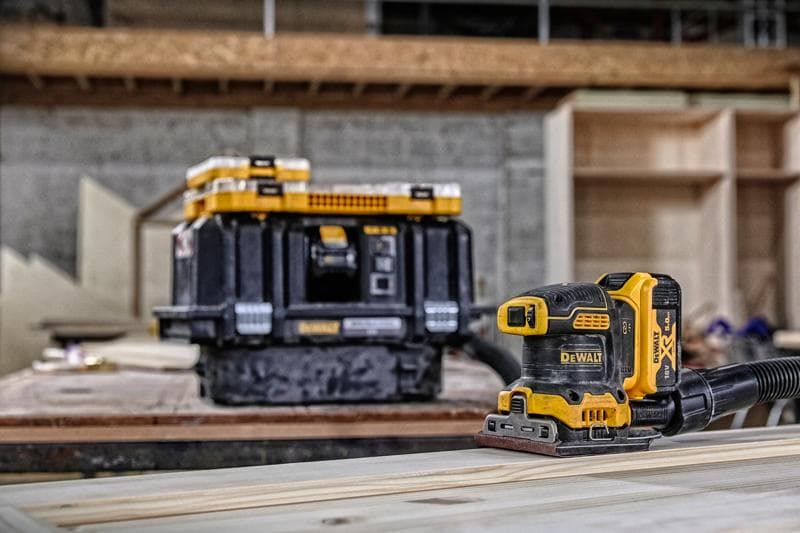 DEWALT Akku-Schleifer auf Holz, mit Aufbewahrungsbox im Hintergrund in einer Werkstatt.