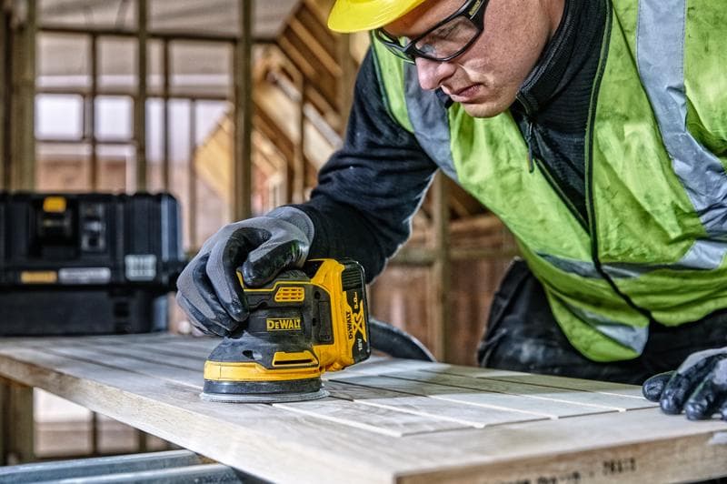 DEWALT DCW210, DCW210D1 Schleifer im Einsatz auf Holz, Baustelle, Person mit Handschuhen und Sicherheitsweste.