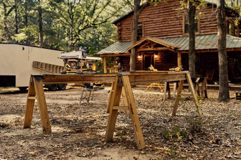 Eine DEWALT Tischkreissäge steht auf einer Holzwerkbank aus zwei Sägeböcken im Freien, im Hintergrund eine Blockhütte und Bäume.